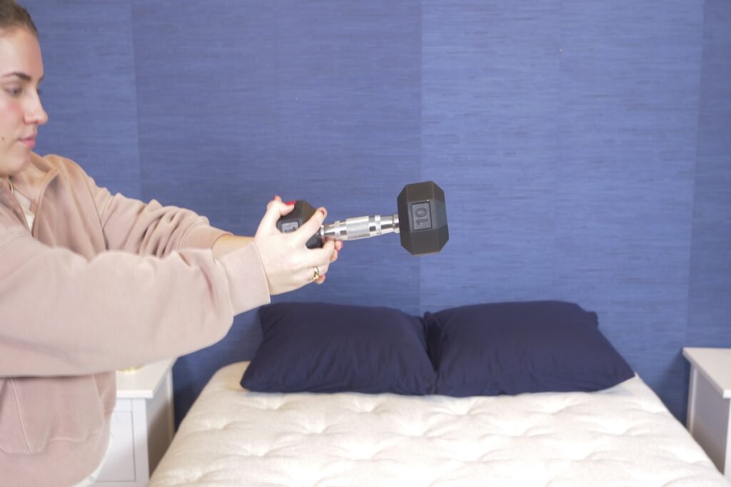 A woman holds a 10-pound dumbbell over a mattress with blue pillows